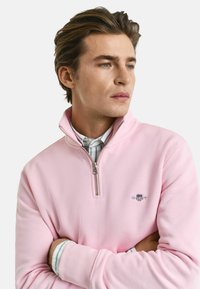 Pullover zip-up rosa chiaro in tessuto morbido, con colletto alla mandarino e logo ricamato. Indossato sopra una camicia a righe con colletto.