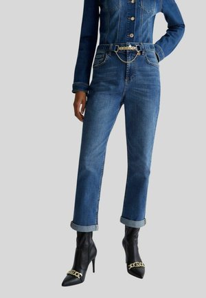 Woman wearing rolled-up blue jeans, denim jacket, black heeled ankle boots with gold chain detail, standing against gray background.