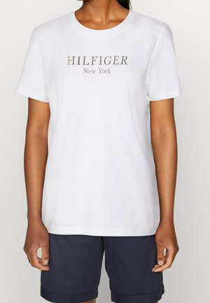 Personne portant un t-shirt blanc à manches courtes avec le texte "HILFIGER New York" et un short bleu marine foncé, debout devant un fond uni.