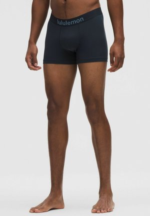 Schwarze, eng anliegende Herren-Boxershorts mit gebrandetem Bund, getragen von einem dunkelhäutigen, stehenden Modell vor neutralem Hintergrund.