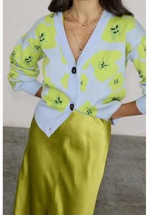 Femme portant un cardigan bleu avec de grands motifs floraux jaune-vert, des boutons noirs, des colliers en or superposés et une jupe en satin jaune-vert brillante.