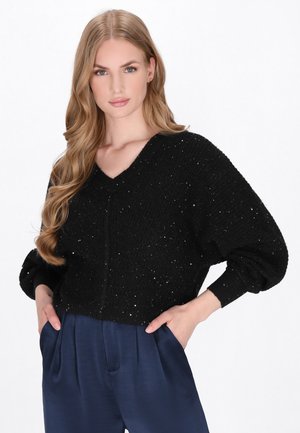Pulover negru cu guler în V, cu un model de tricotat texturat, având străluciri argintii. Mânecele sunt largi și se îngustează la încheieturi.