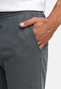 Szare, teksturowane spodnie z gładkim wykończeniem, z kieszeniami bocznymi i ręką opartą w jednej z kieszeni, podkreślając wzór splotu materiału.