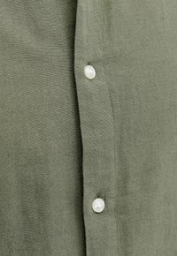 Nærbillede af olivengrønt stof med to hvide knapper syet lodret på en skjorteliste.