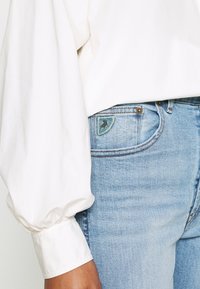 Person in hellblauen Jeans mit einem kleinen Pferde-Patch an der Tasche und einer weißen Bluse mit langen Ärmeln und gepufften Manschetten.