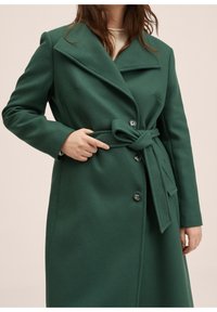 Grøn, lang frakke i uldblanding med bredt revers, bindebælte, enkeltradet front og knapdetaljer, vist på en person med den ene hånd på bæltet.