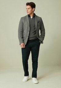 Grau Blazer über einem gemusterten dunklen Pullover, kombiniert mit dunklen Hosen und weißen Sneakern. Ein Einstecktuch sorgt für einen Farbtupfer.