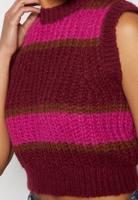 Gros plan sur un gilet sans manches en tricot avec de larges rayures horizontales bordeaux, roses et marron sur le torse d'une personne.
