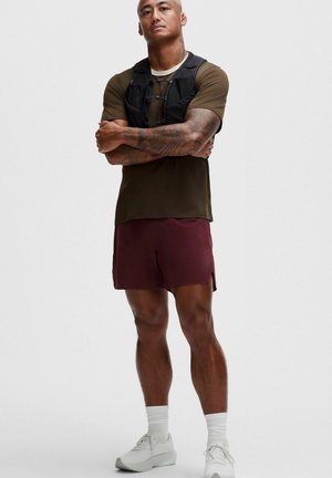 Muskelbepackter Mann mit Tätowierungen steht mit verschränkten Armen da, trägt ein olivgrünes Hemd, schwarzes Brustgeschirr, dunkelrote Shorts, weiße Socken und weiße Turnschuhe.