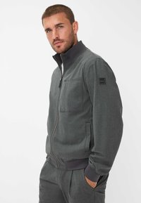 Mann mit kurzem Haar und Bart, der eine graue Zip-Jacke und passende Hose trägt, steht mit einer Hand in der Tasche vor einem weißen Hintergrund.