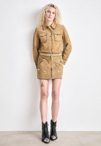 Giacca in suede color tan con due tasche frontali e cuciture nere a contrasto, abbinata a una gonna coordinata con dettagli di zip e borchie. Stivaletti neri alla caviglia con punta argentata.