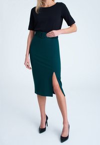 Žena v černém krátkém rukávu topu zastrčeném do tmavě zelené tužkové sukně s vysokým pasem, páskem a postranními rozparky, černé vysoké podpatky.