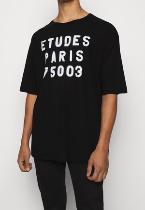 Černé bavlněné tričko s oversized střihem, kulatým výstřihem a bílým grafickým textem na přední straně: "ETUDES PARIS 75003." Krátké rukávy s uvolněným siluetou.