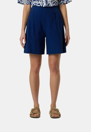 Donna che indossa pantaloncini plissettati blu navy a vita alta con tasche laterali, sandali beige e una camicia fantasia blu e bianca.