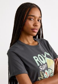 T-shirt gris à manches courtes avec un motif floral imprimé et le texte "ROXY SURF". Matière douce avec un col rond et des manches à revers.