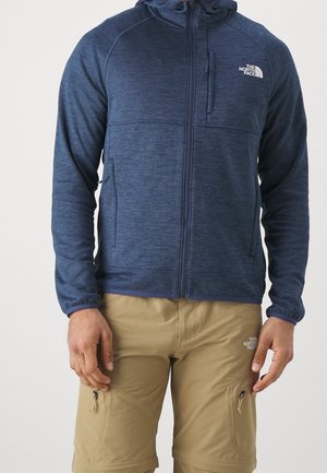 Mężczyzna ubrany w niebieską kurtkę z kapturem zapinaną na zamek oraz beżowe szorty, oba z logo The North Face, stojący na jednolitym tle.