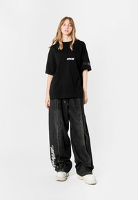 Camiseta negra de gran tamaño con el logotipo "MWM", combinada con pantalones de mezclilla negros holgados que tienen franjas laterales y acentos blancos. Calzado de color claro.