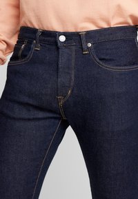 Mörkblå denimjeans med kontrasterande orange sömmar, bärs med en ljus persikofärgad topp, med fokus på midje- och övre lårområdet.