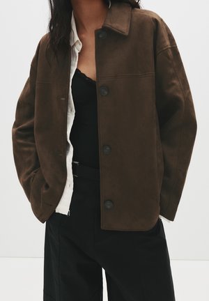 Person wearing a dark brown buttoned jacket over a white shirt and black top, with black pants, standing with hands in pockets.