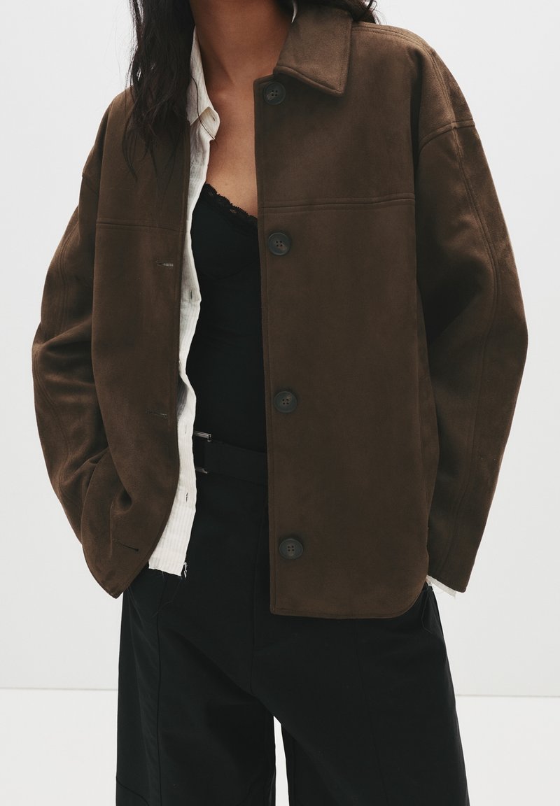 Personne portant une veste marron foncé boutonnée sur une chemise blanche et un haut noir, avec un pantalon noir, debout les mains dans les poches.
