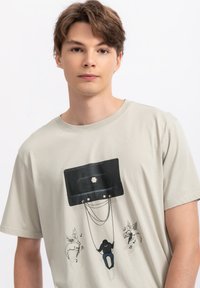 Lichtbeige katoenen T-shirt met een zwart cassette-ontwerp waarop een figuur op een schommel staat, omringd door gevleugelde paarden en muzieknoten.
