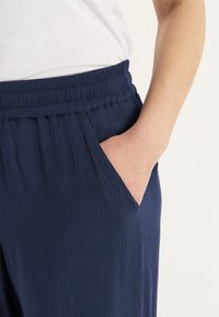 Marineblauwe broeken gemaakt van een gestructureerde, gekreukte stof. Voorzien van een elastische tailleband en een zijzak, getoond van de taille naar beneden.