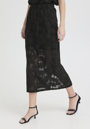 Kvinde iført en sort midinederdel med blomsterblonde og sorte åbne tå-hælstropper, stående mod en ensfarvet baggrund.