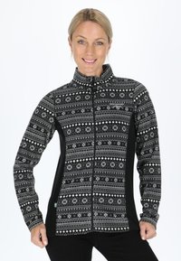 Smiling woman wearing a black and white patterned zip-up fleece jacket, standing with hands at hips against a white background.