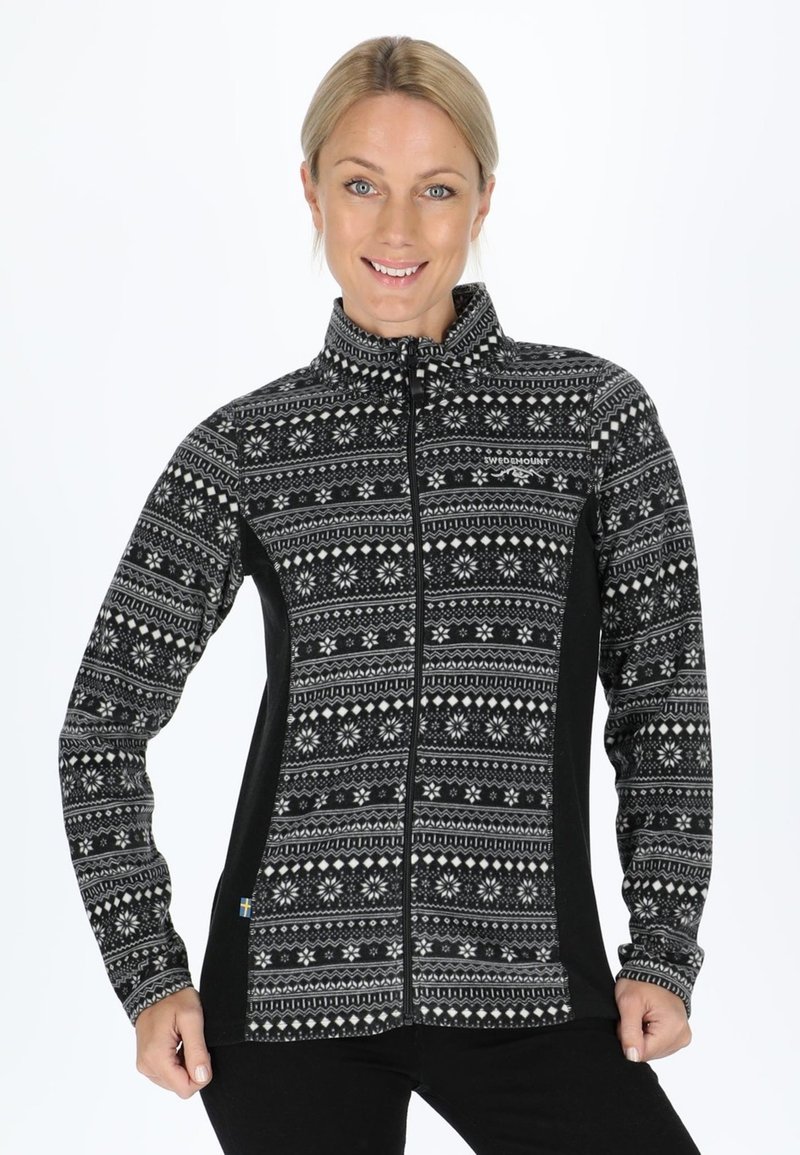 Smiling woman wearing a black and white patterned zip-up fleece jacket, standing with hands at hips against a white background.