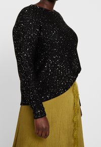 Maglione nero con paillettes metalliche, scollo rotondo e maniche lunghe, abbinato a una gonna gialla plissettata con bordatura in pizzo.