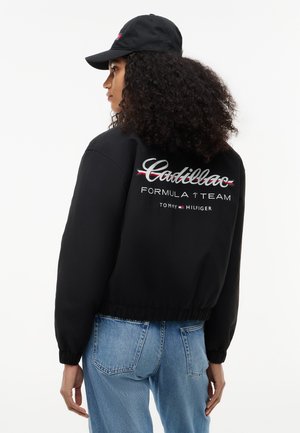 Mujer con cabello rizado lleva una gorra negra y chaqueta con el logo "Cadillac Formula 1 Team Tommy Hilfiger", combinada con vaqueros azules.