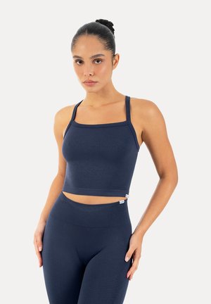 Frau mit zusammengebundenen Haaren im Dutt, trägt ein marineblaues bauchfreies Tanktop und hoch taillierte Leggings, steht vor weißem Hintergrund.