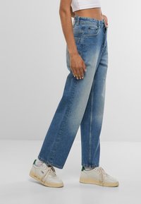 Gaiši zili augsta vidukļa džinsi ar brīvu piegriezumu, kuriem ir piecas kabatas, izbalējuma detaļas un rāvējslēdzēja aizdare. Apvienoti ar baltiem sporta apaviem.