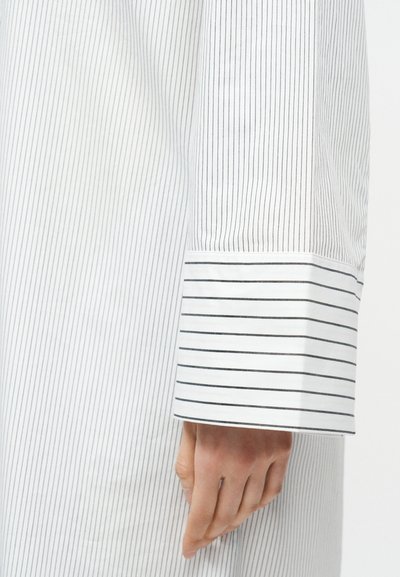 Κοντινό πλάνο ενός χεριού που ακουμπάει δίπλα σε λευκό μανίκι με λεπτές κάθετες μαύρες ρίγες και ευρεία μανσέτα με οριζόντιες ρίγες.