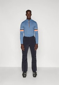 Άνδρας που στέκεται σε μπλε μακρυμάνικο μπλουζάκι με ρίγες στα μανίκια, μαύρα παντελόνια και μπότες σκι μπροστά από λευκό φόντο.