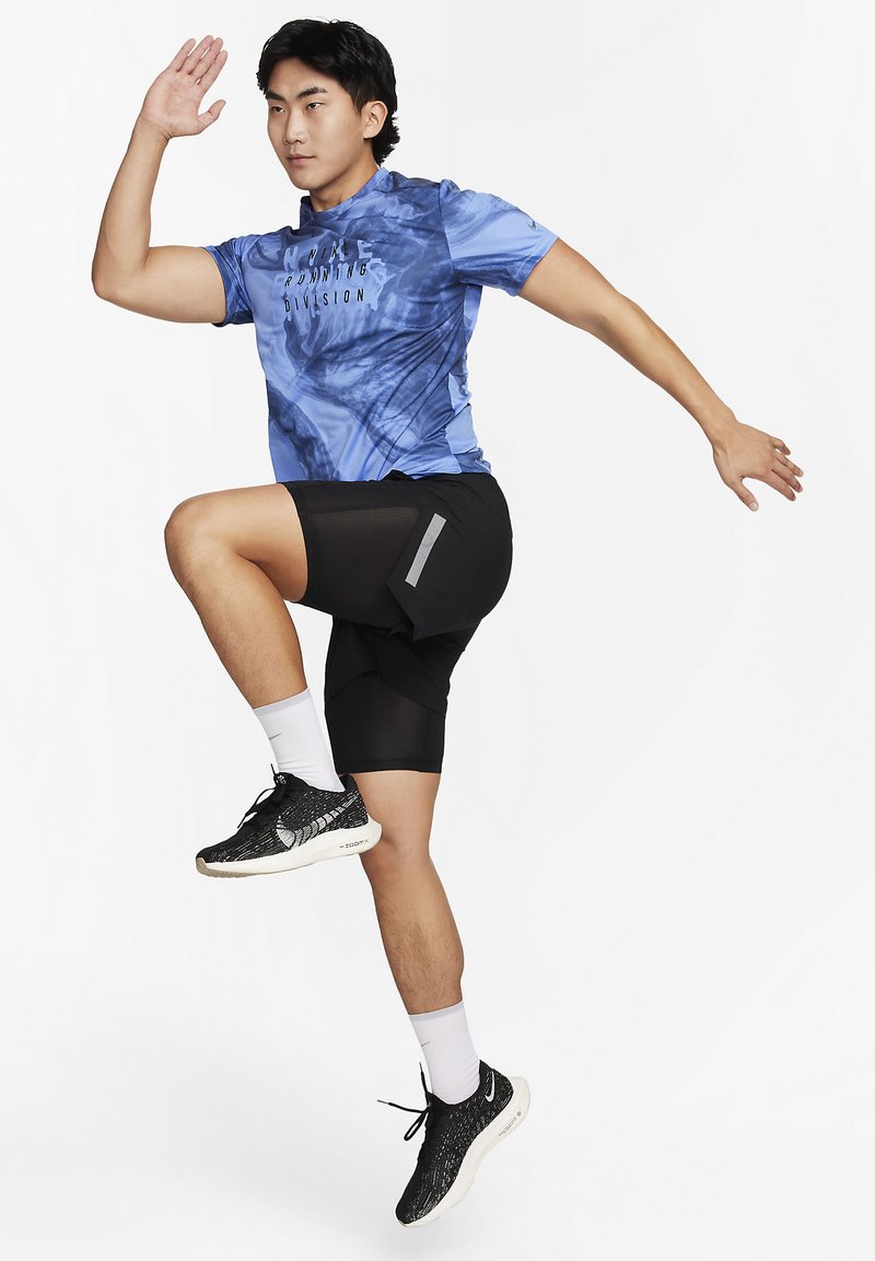 T-shirt de course bleu avec un design abstrait, shorts noirs avec poche latérale et baskets noires texturées. Chaussettes blanches visibles.