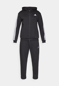 Črni športni trenirka iz mehke tkanine; jopa z zadrgo in belimi črtami na rokavih ter logotipom na prsih; ujemajoče ozke hlače z logotipom.