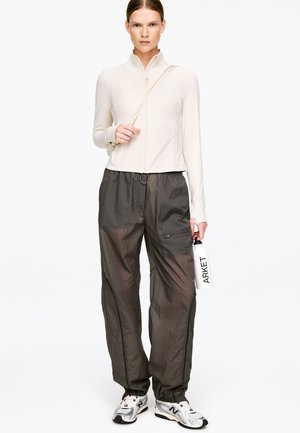 Lekka beżowa kurtka zapinana na zamek z wysokim kołnierzem, zestawiona z luźnymi ciemnoszarymi spodniami z kieszeniami. Model trzyma białą butelkę na wodę.