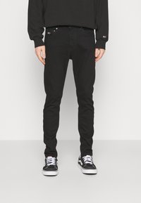 Černé slim-fit džíny s pěti kapsami a kovovými doplňky, spárované s černými teniskami s bílými tkaničkami a značkováním.