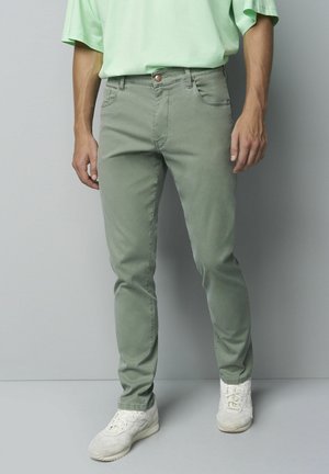 Man wearing slim-fit sage green jeans, light green t-shirt, and white sneakers standing against a gray background.