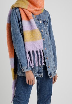 Personne portant une veste en jean bleue et un jean avec une grande écharpe duveteuse rayée orange, lavande et jaune enroulée autour du cou.