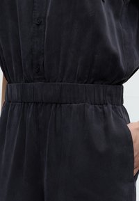 Κοντινό πλάνο ενός ατόμου που φοράει μαύρο πουκάμισο με κουμπιά και αντίστοιχο παντελόνι με ελαστική μέση, το χέρι ακουμπάει στην τσέπη της δεξιάς πλευράς.