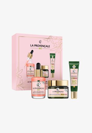 Ensemble de produits de soin de la peau dans une boîte rose. Comprend des bouteilles en verre avec des accents dorés, un pot de crème foncé et un tube vert. Différentes étiquettes visibles.