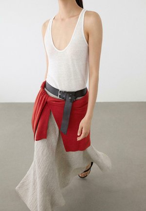 Woman in white sleeveless top, grey flowing skirt, black belt, red sweater tied at waist, walking on a smooth grey floor wearing black sandals.