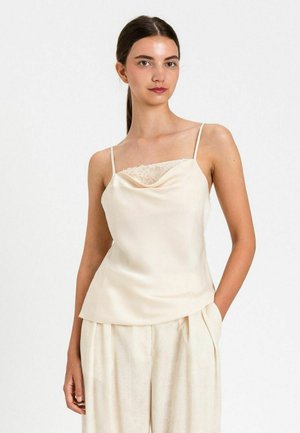 Donna che indossa una canottiera in satin crema con dettaglio in pizzo e pantaloni larghi abbinati, in piedi con una mano in tasca su sfondo bianco.