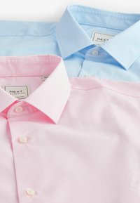 Dos camisas de vestir con cuello, una de color azul claro y otra de color rosa claro, dispuestas planas, con una textura suave y botones blancos. Ambas tienen un corte ajustado.