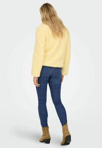 Yellow faux fur jacket with a textured finish, oversized sleeves, and a cropped design, paired with dark blue skinny jeans and tan ankle boots.