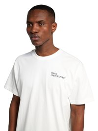 Mężczyzna ubrany w białą koszulkę z napisem „HALO 196819721981” małym czarnym tekstem na lewej piersi, stoi na białym tle.