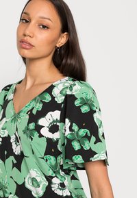 Blusa preta com padrões botânicos verdes e brancos, decote em V e mangas curtas soltas com uma textura suave.