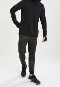 Musta pitkähihainen huppari, jossa on peukalonreiät, yhdistettynä tummanvihreisiin maastokuvioisiin verkkareihin ja mustiin urheilusandaaleihin. Yksinkertainen ja toiminnallinen muotoilu.